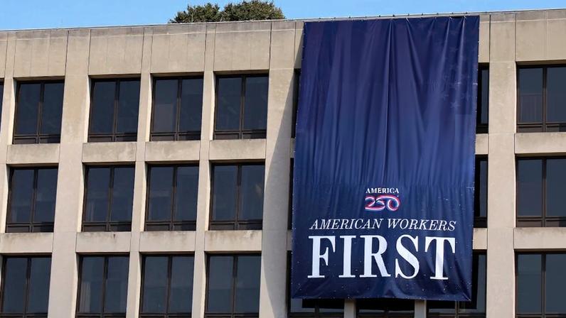 A massive blue banner with Donald Trump's photo and the words "American Workers First" hangs from a brutalist-style building. The banner covers up at least six of the building's windows.