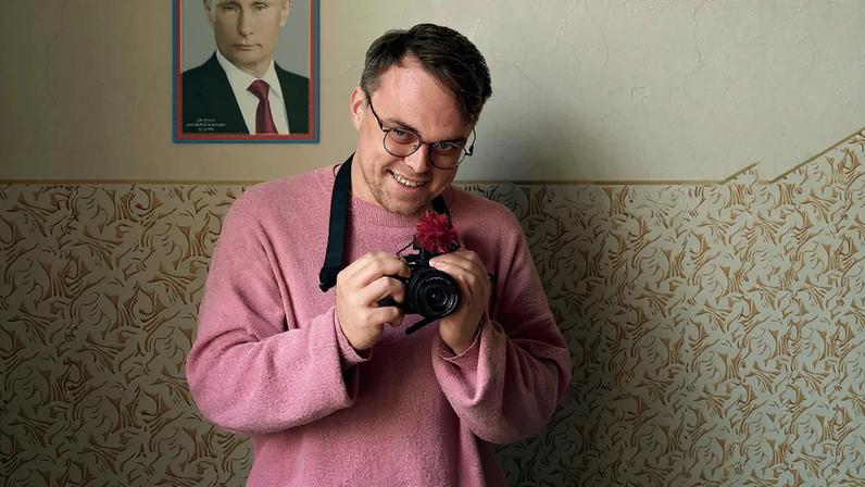 A man in a pink shirt, holding a camera, smiles in front of a poster of Putin.