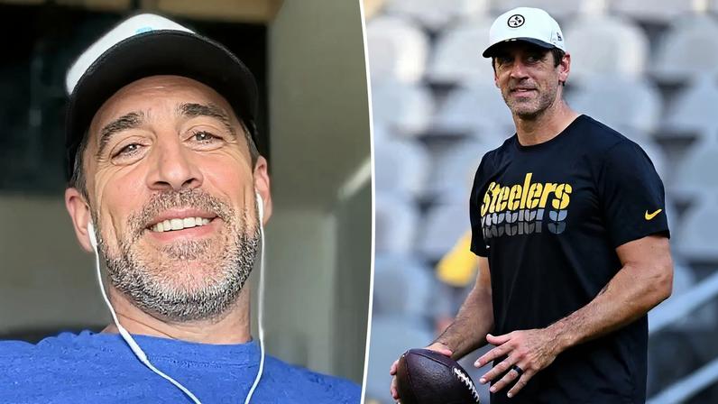 Aaron Rodgers smiling while wearing a cap and wired earbuds.