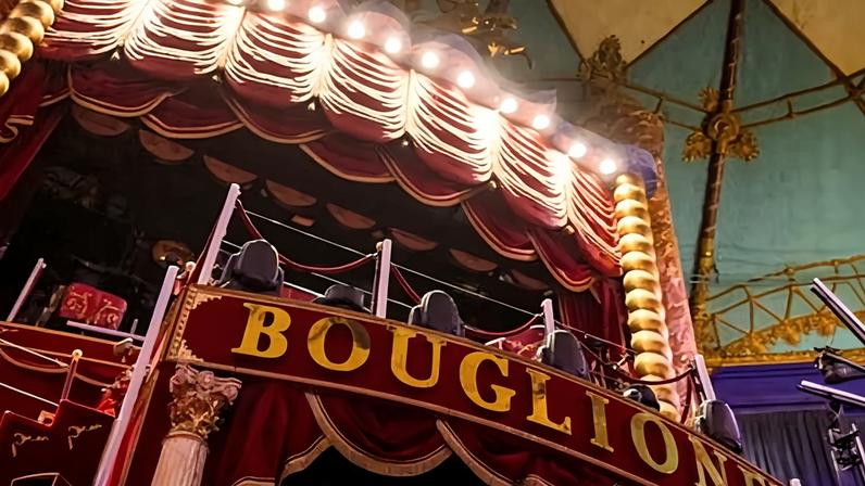 Interior of the Cirque d’Hiver in Paris, showing its circular ring surrounded by tiers of red seats and an ornate 19th-century balcony beneath a domed roof with a sign reading 'Bouglione'.