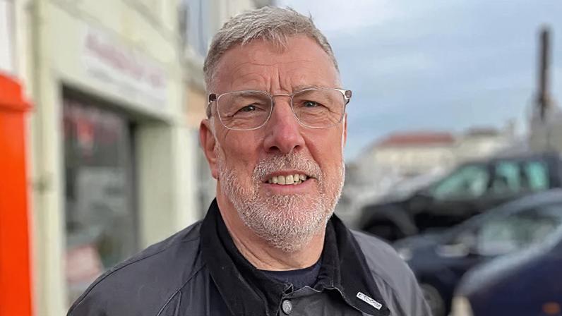 Deputy Adrian Gabriel from the States of Guernsey stood outside some shops and parked cars. He is looking straight at the camera and is smiling.