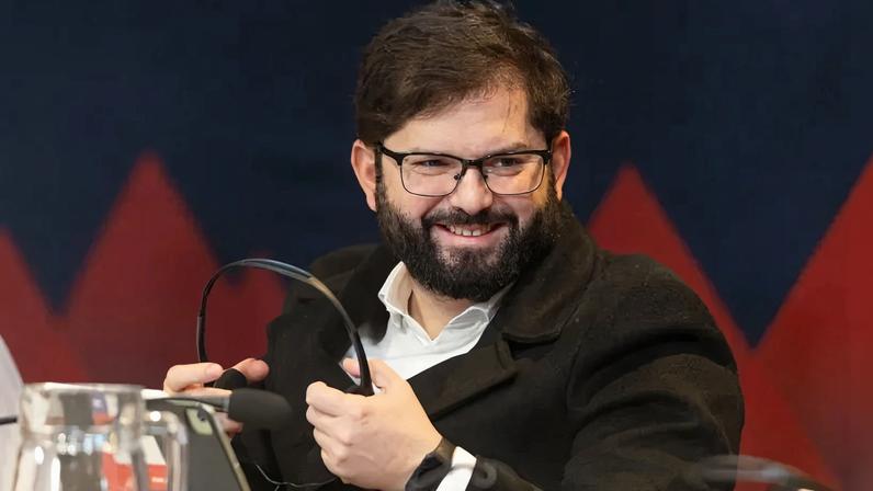 Boric smiles as he holds a pair of headphones. He is a man in his late 30s wearing a white shirt and black coat in front of a blue and red background.