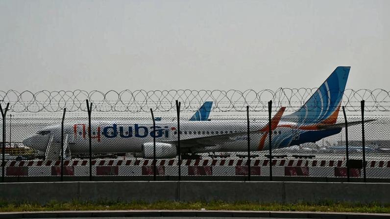 A FlyDubai airplane with blue, white, and orange designs is parked behind a barbed wire fence at Dubai International Airport.
