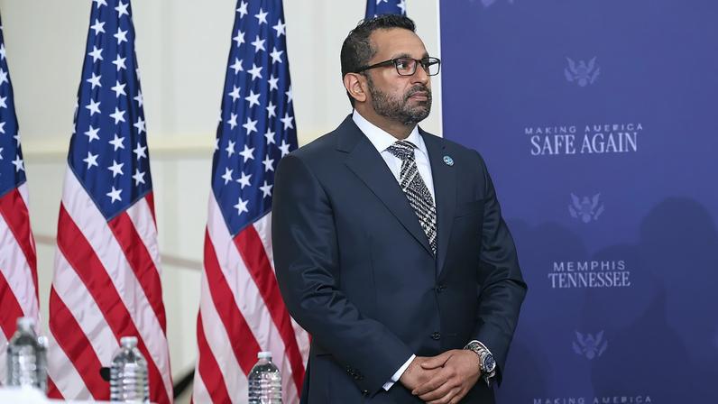 Kash Patel stands with his hands clasped in front of several U.S. flags.