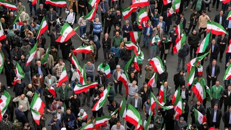 A crowd of dozens of people, mostly but not all men, is seen from above as they wave Iranian flags on poles.