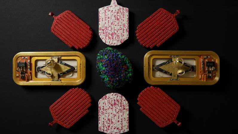 The device and pieces are arranged in a mandala pattern. The device has liquid inside and bubbles. The pieces have pathways, some filled with pink dots.