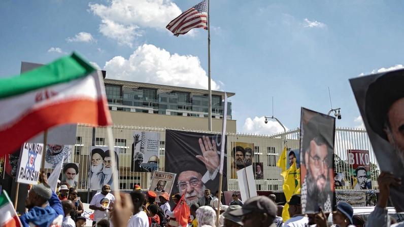People are seen outside the U.S. consulate, with a U.S. flag flying above it. On the fence along the consulate are posters of Iranian leaders.