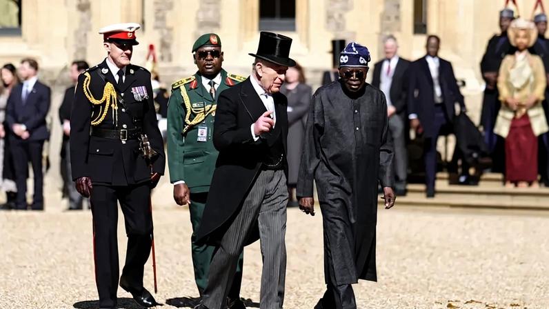 King Charles III and Bola Tinubu walk side by side; a small crowd of people stands behind them.