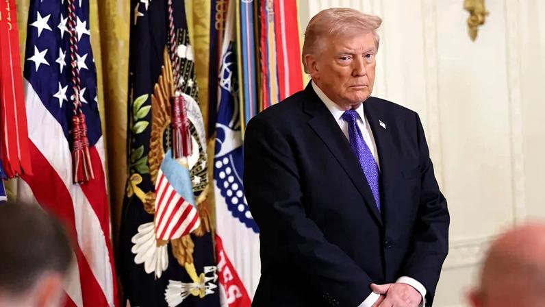 U.S. President Donald Trump stands in front of U.S. flags with his hands clasped in the East Room of the White House.