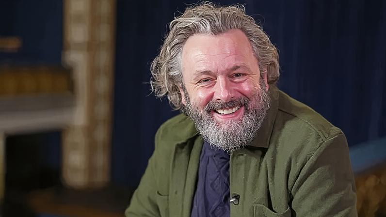 Michael Sheen sits in a theatre looking at the camera. He has long wavy greying hair and a beard. He is wearing a green jacket and blue jumper.