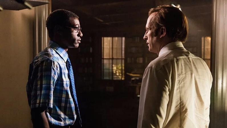 Two men, one in a blue plaid shirt and blue tie and the other in a white shirt, face each other in a dimly lit room.