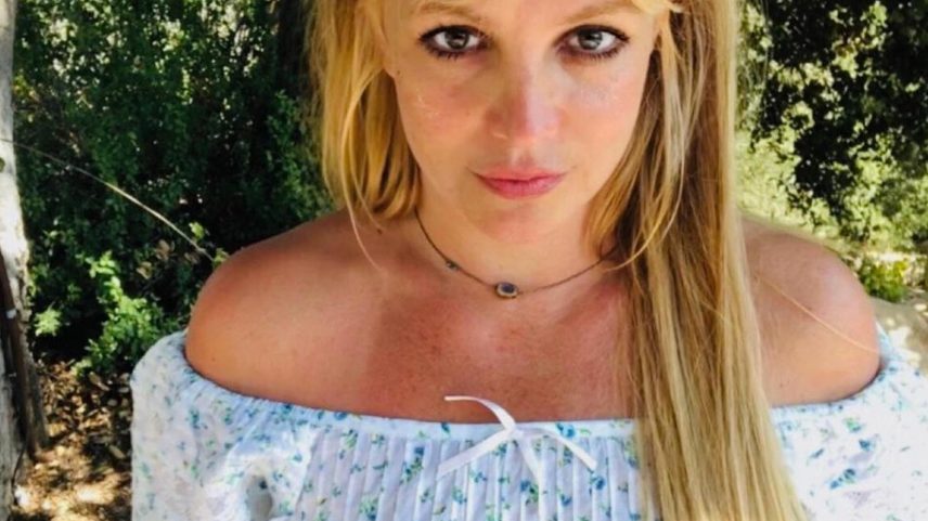 Britney Spears with long blonde hair, wearing a white off-the-shoulder crop top with blue floral print.