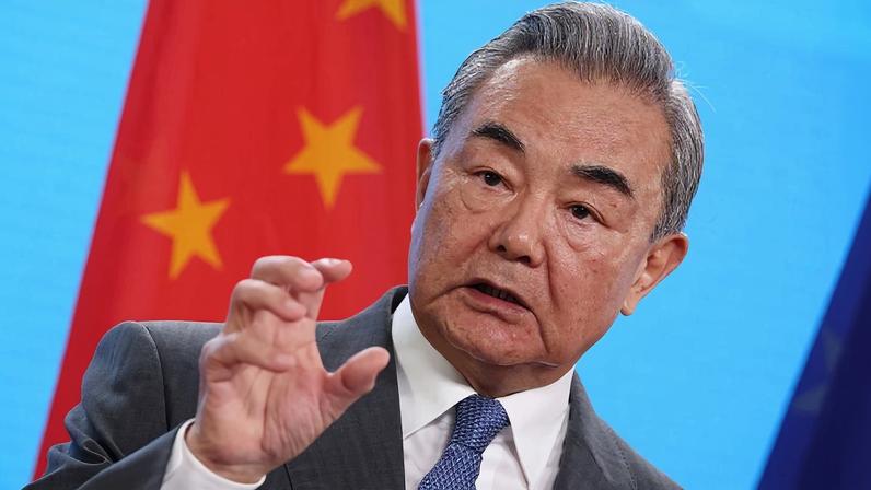 Wang, wearing a gray suit and tie, gestures with one hand bent into a claw-like shape while speaking in front of a Chinese flag and a bright blue background.