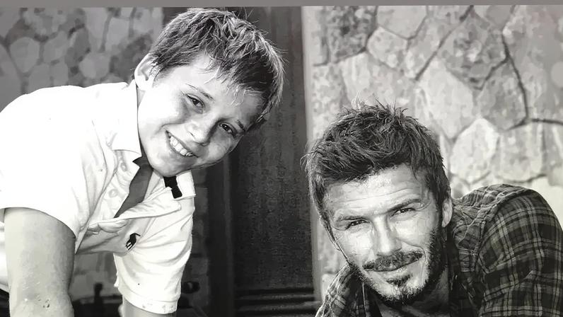 David Beckham and a young Brooklyn Beckham smiling and leaning over a barrel.