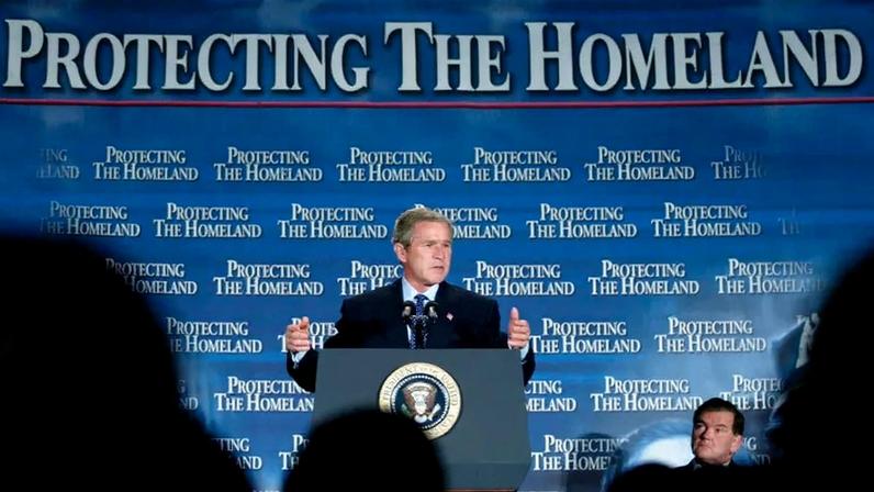 George W. Bush gestures as he speaks behind a lectern in front of a backdrop with the words "Protecting the Homeland" repeated behind him.