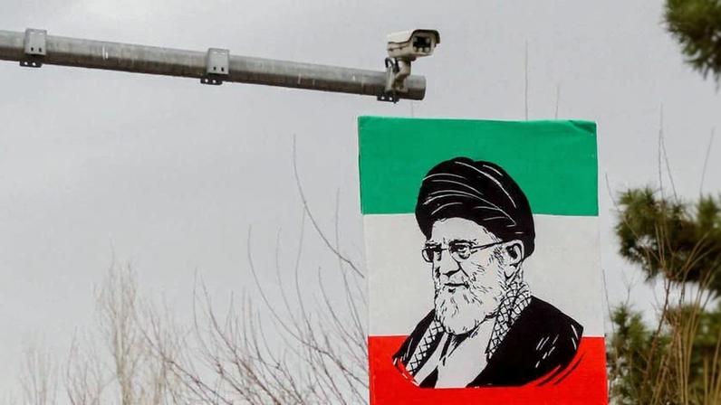 A poster featuring a black-and-white drawing of Ali Khamenei against a green, red, and white Iranian flag-style background is seen near a traffic camera.