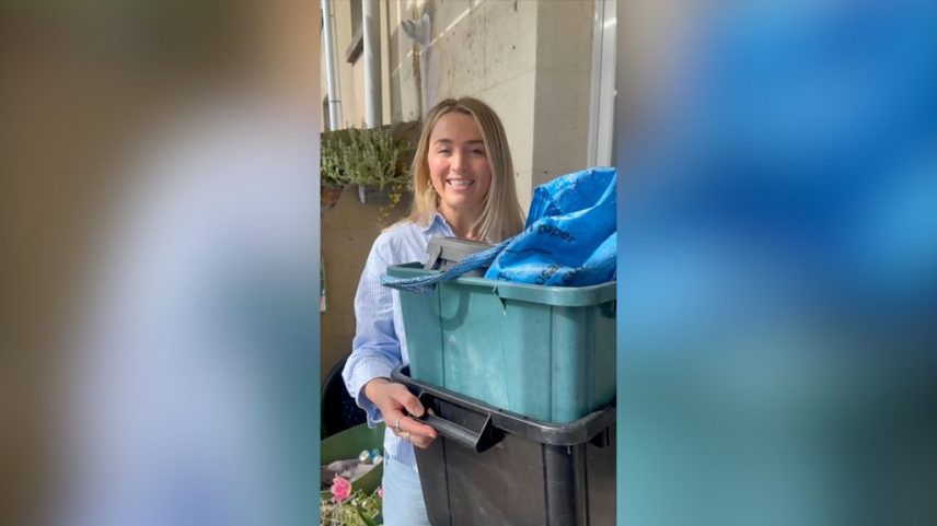 A woman with long blonde hair talks to the camera while holding two recycling tubs