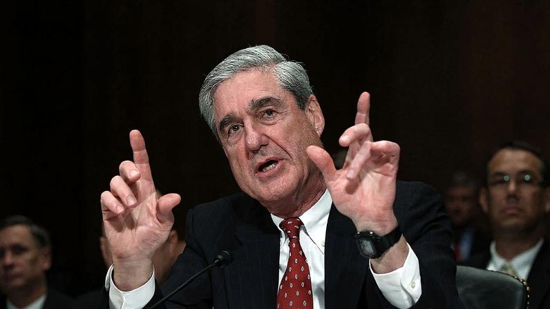 Robert Mueller gestures with both hands as he talks in front of a microphone.