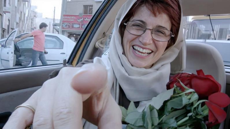 A woman in a headscarf holding a bunch of red roses smiles andn points at the camera from the back of a taxi.