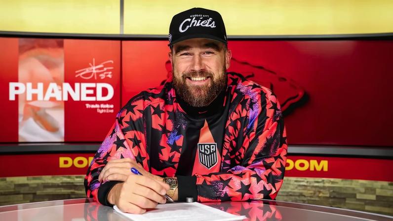 Travis Kelce signing a contract, wearing a USA jersey and a Kansas City Chiefs cap.