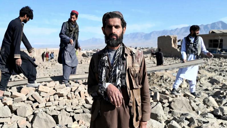 A bearded man with a bandage high up on his cheek and one arm in a makeshift sling made from a scarf stares at the camera. Behind him, three other men climb around a pile of rubble, seemingly mostly stone and concrete, from a demolished building.
