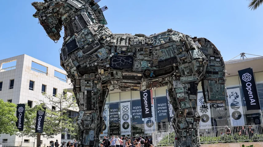 A giant horse looms over an outdoor plaza in front of a building about four stories tall. The horse is constructed of pieces of scrap metal and tech equipment, such as computer monitors, keyboards, and circuit boards.