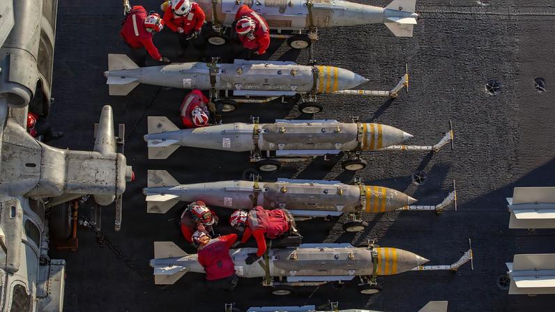 About half a dozen U.S. Navy sailors are seen from overhead as they work on what appear to be torpedos or missiles of some sort. The sailors wear bright red uniforms and helmets.