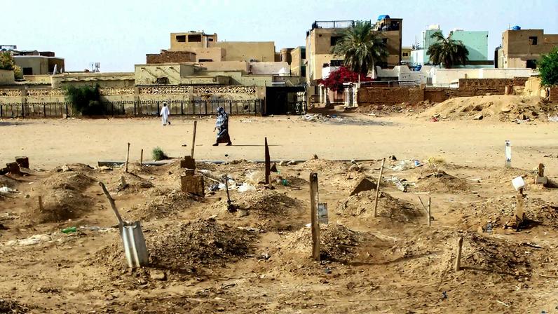 In a landscape almost entirely covered with light brown sand, a small cemetery in the foreground is filled with mounds of dirt marked with sticks or small stones. Beyond that, a woman in an ankle-length dress and a headscarf walks by, her shadow long behind her. Low-rise buildings in a similar sandy color are visible beyond a fence behind her.