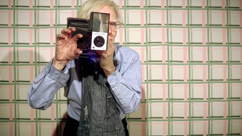 Andy Warhol in an apron with his Big Shot Polaroid camera in the Factory (1970s) by Ronnie Cutrone
