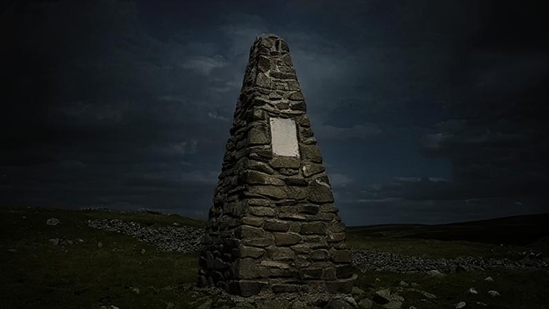 Walter Baxter A pyramid shaped stone cairn on the top of a Borders hillside. There are stones littered around it on a grassy landscape with a cloudy blue sky above.
