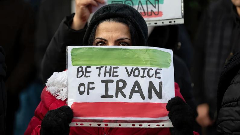 A close-up shot of a person wearing a black head covering and a red jacket, holding up a handmade sign that covers the lower half of their face. The sign features green and red brushstrokes at the top and bottom to resemble a flag, with the handwritten text "BE THE VOICE OF IRAN" in bold black letters.