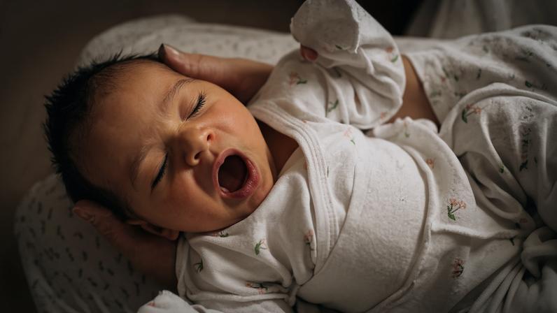 Bebê recém-nascido deitado de costas, bocejando com a boca aberta e os olhos fechados, vestido com um macacão branco estampado com pequenas flores cor-de-rosa, sendo segurado por mãos adultas em um fundo claro