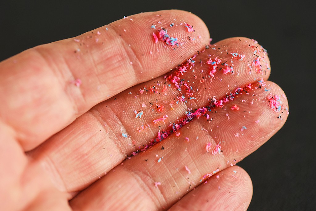 Mão aberta com fragmentos coloridos de plástico rosa, azul e preto espalhados sobre a palma e os dedos, em fundo escuro