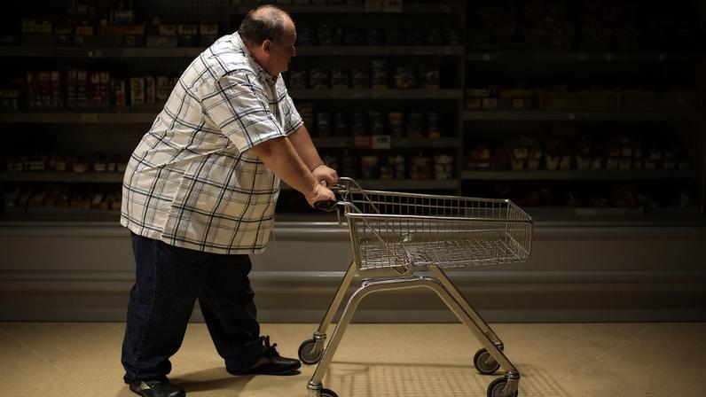 Homem de meia-idade, com sobrepeso, camisa xadrez e calça jeans, empurra um carrinho de compras vazio em um supermercado, passando por prateleiras de frutas, legumes e produtos enlatados