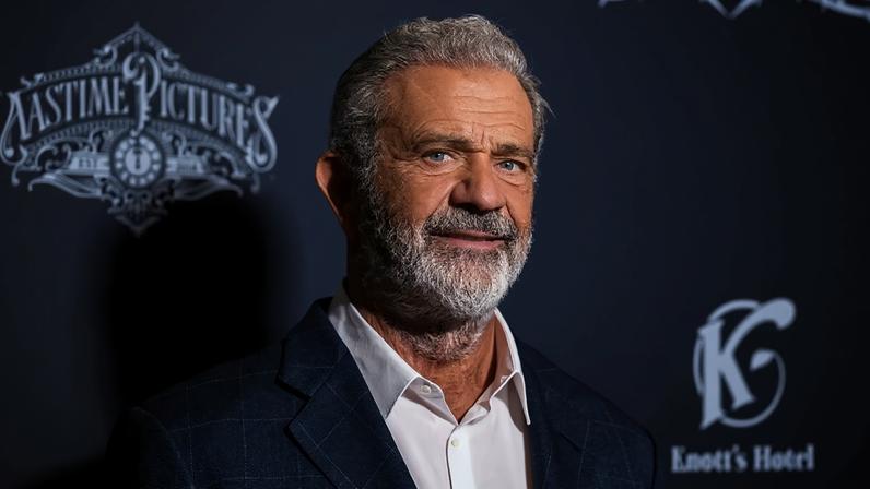 Mel Gibson, homem branco de cabelos grisalhos e barba, sorrindo levemente, vestindo blazer xadrez azul e camisa branca, em frente a um fundo escuro com logotipos