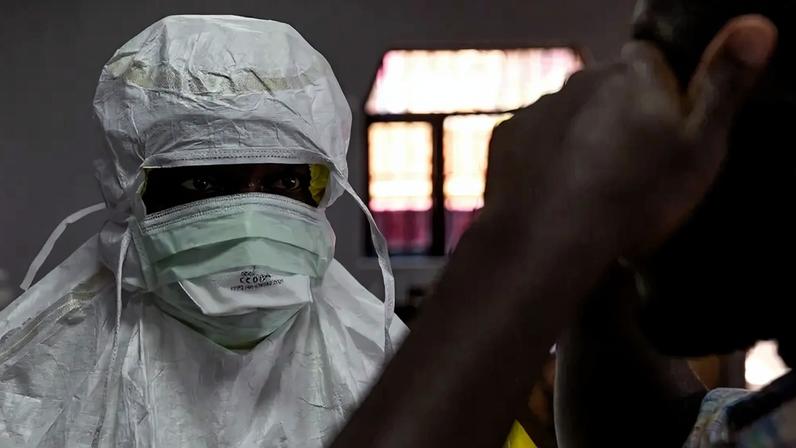 Fotografia de um homem negro que é Profissional de saúde com EPI branco e máscara verpe, olhando fixamente, com um paciente de pele escura em primeiro plano desfocado à direita