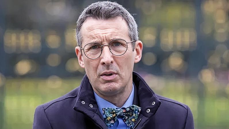 PA Media Andrew Muir is standing outside on front of large black gates. He wears glasses and is wearing a navy coat with a blue shirt and a bow tie.