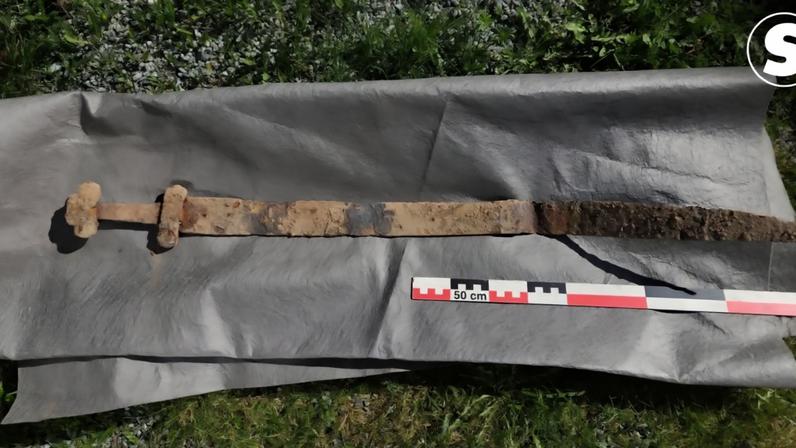 Espada antiga e enferrujada, com cabo em forma de T, sobre um tecido cinza escuro, com uma régua de 50 cm para escala, em um gramado com pedras