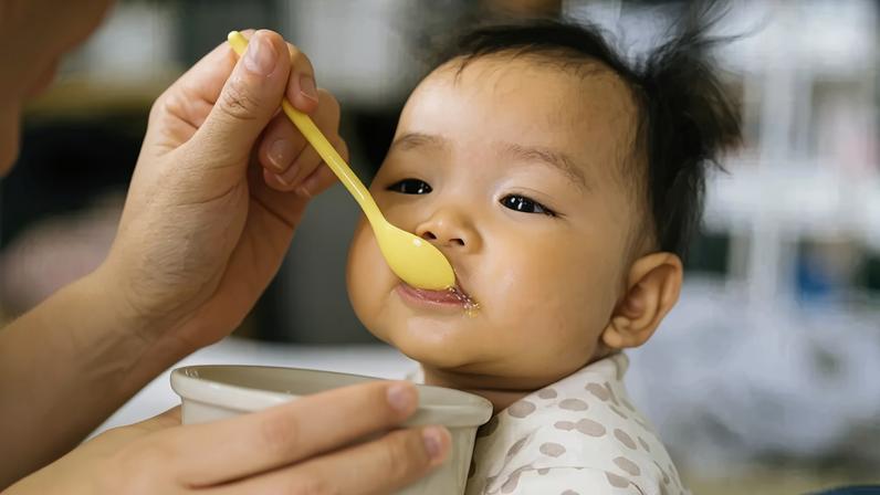 Mão adulta alimentando um bebê asiático com uma colher amarela, o bebê usa um macacão claro com bolinhas e tem um pouco de comida na boca