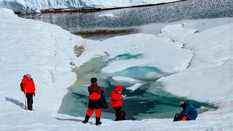 Quatro pessoas em roupas de inverno, três de casaco vermelho e uma de casaco azul, observam um lago de água azul-esverdeada cercado por neve e gelo. Uma pessoa à esquerda segura um bastão, duas ao centro olham para a água e uma à direita está agachada. Ao fundo, uma grande parede de gelo e um lago maior.