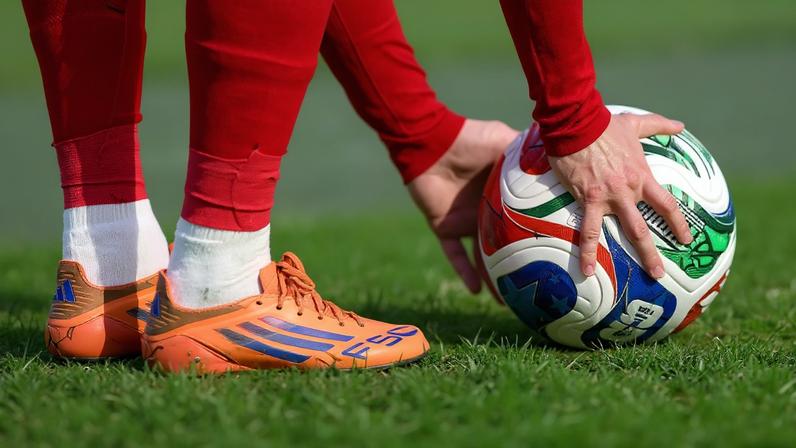Um jogador de futebol, vestindo uniforme vermelho e chuteiras laranja com detalhes azuis, posiciona uma bola colorida no gramado verde, próximo à linha branca do campo