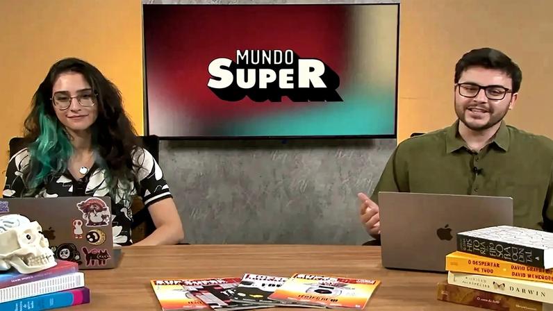 Dois jovens, uma mulher e um homem, sentados em uma mesa de madeira com laptops, livros e revistas. A mulher tem cabelo escuro com mechas azuis e usa óculos e blusa estampada. O homem usa óculos e camisa verde-oliva, gesticulando. Ao fundo, uma tela exibe o logo "Mundo Super"