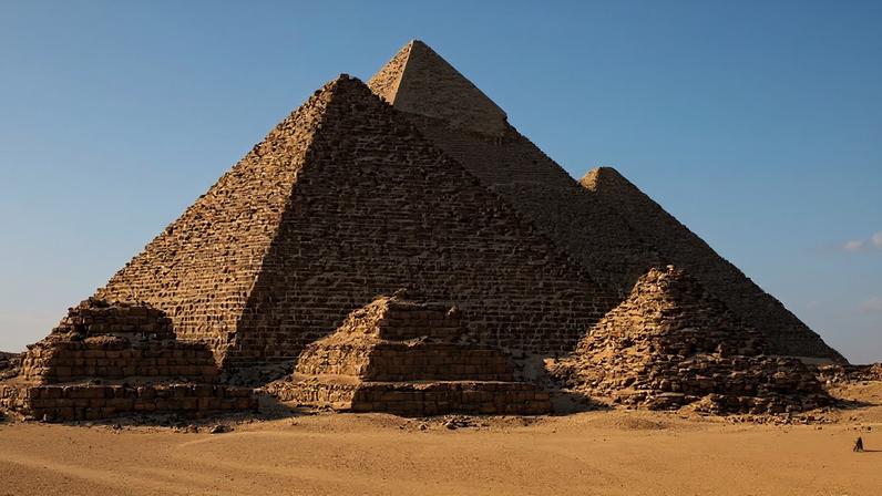 Três pirâmides de Gizé, no Egito, sob um céu azul claro, com a maior em primeiro plano e duas menores ao fundo. A areia dourada do deserto se estende à frente, e dois camelos com cavaleiros são visíveis à direita