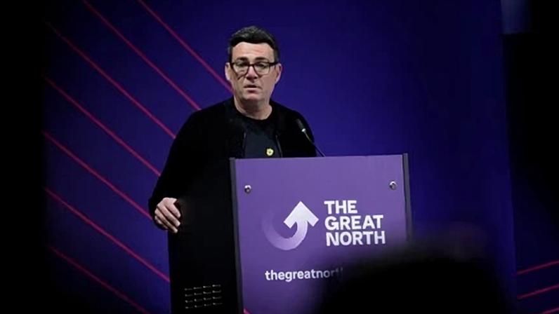 Andy Burnham speaks at an event. He stands illuminated on a dark stage with shadows around him and in front of a purple backdrop with red stripes. A sign on his lectern has an arrow logo and reads The Great North. He wears black.