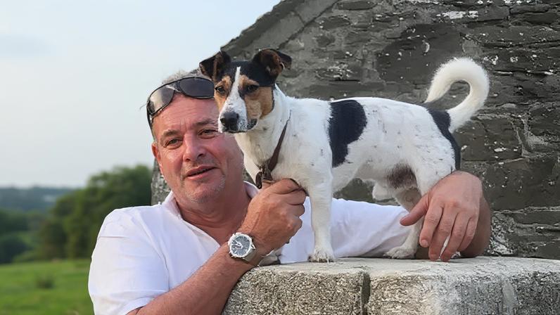 James Southey looking and smiling at the camera with his small dog stood on a wall.