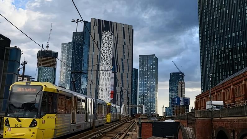 A yellow tram travels along tracks in Manchester with modern glass skyscrapers rising behind under cloudy sky