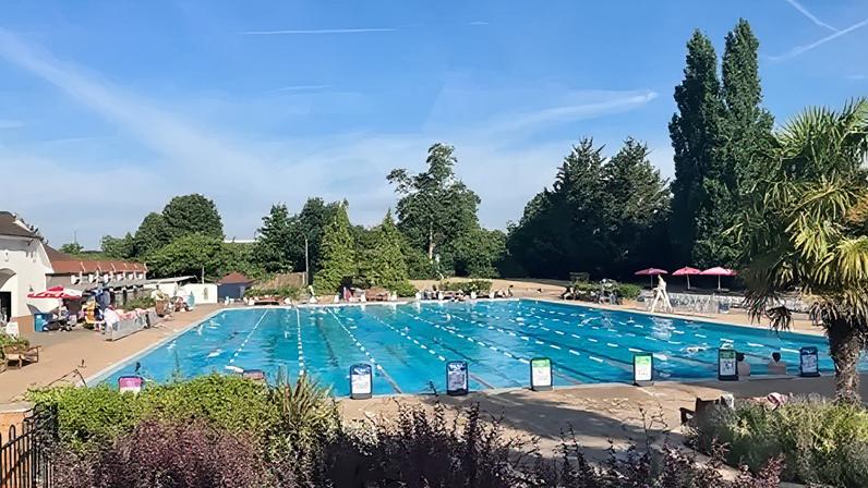 Guildford Lido, an outdoor pool, is seen under a blue sky and it is surrounded by trees. The water is blue and there are lane dividers out and some swimmers swimming lengths.