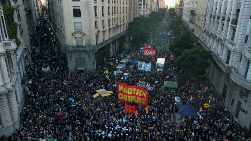 Thousands of people crowd in an intersection between two city streets, with the crowds stretching down the streets beyond view. Many hold banners or smaller signs.
