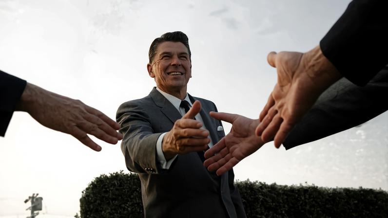 A black and white photograph from a low angle shows a smiling man in a suit reaching out to shake hands with several people. His arm is extended toward the camera, and the hands of three other people in suits enter the frame from the sides, reaching back toward him. The background features a clear sky and the top of a manicured hedge.