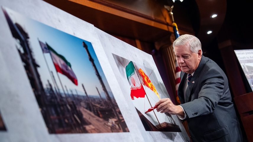 Graham, an elderly man with white hair wearing a business suit, draws with a marker on a photo of an Iranian flag flying outside somewhere.
